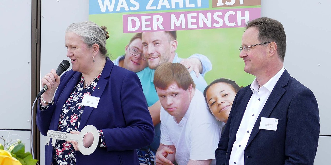 Sabine Ulrich und Thorsten Tillner von der Geschäftsführung der Rotenburger Werke blicken in einer Richtung, Frau Ulrich spricht in ein Mikrofon. Im Hintergrund ein Foto mit jungen Menschen und dem Slogan "Was zählt ist der Mensch"