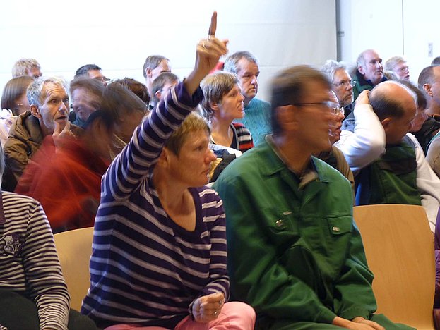 Mehrere Menschen sitzen in Stuhlreihen in einem Saal, eine Frau in der ersten Reihe hat die Hand gehoben