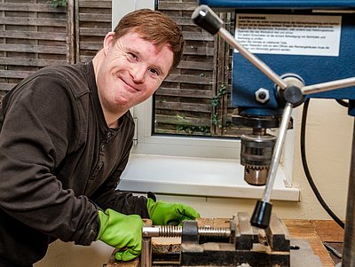 Ein Bewohner arbeitet an einer Werkbank mit Bohrmaschine und lächelt in die Kamera