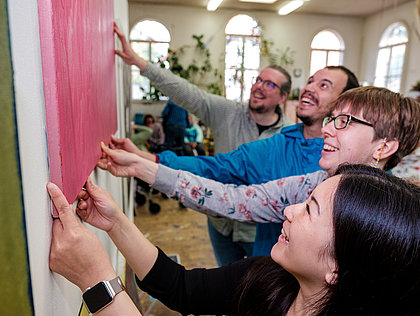 Vier junge Laute  halten in einem Atelier gemeinsam ein Bild in die Höhe