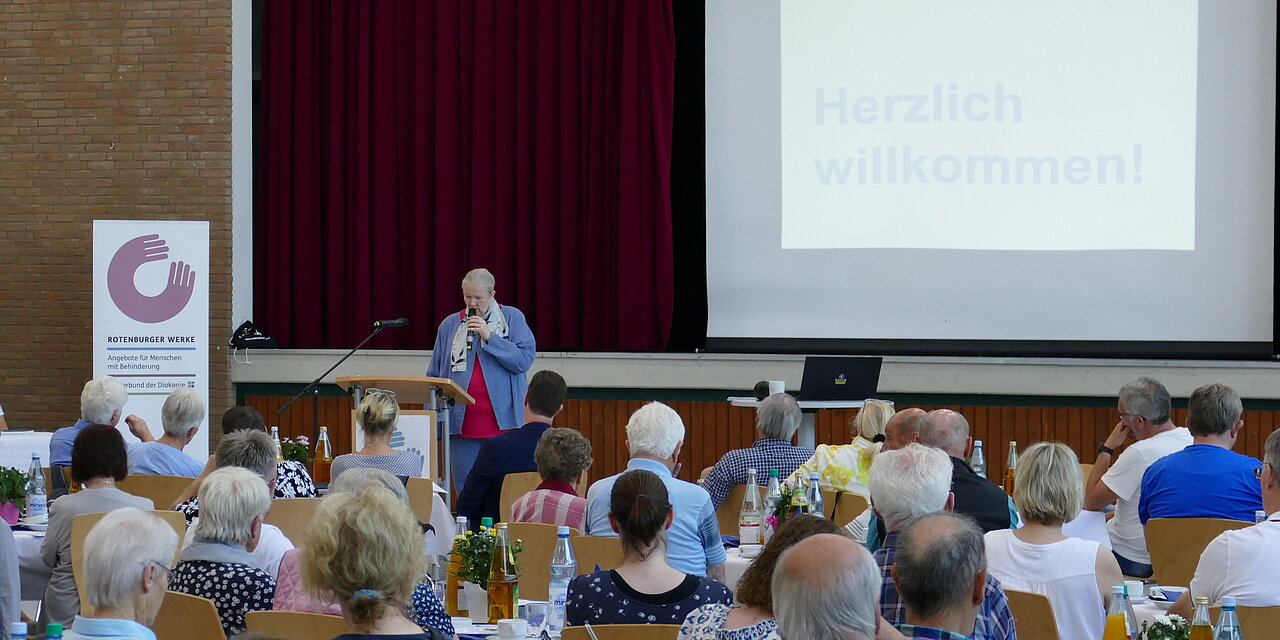 Menschen sitzen in einem Saal an Tischen und lauschen der Worten von Sabine Ulrich, die zu den Gästen spricht
