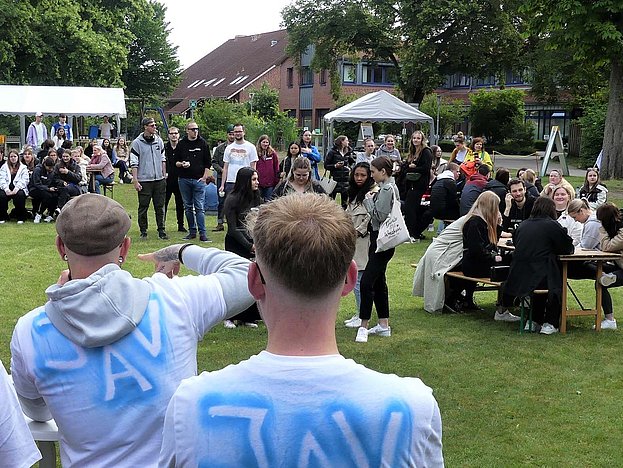 Viele Jugendliche haben sich auf einer Wiese versammelt. Im Vordergrund zwei Männer mit der Aufschrift JAV auf dem Rücken
