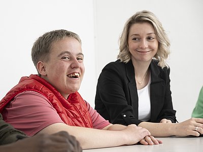 Eine junge Bewohnerin sitzt mit einer Mitarbeiterin am Tisch, beide sind im Gespräch und lächeln.