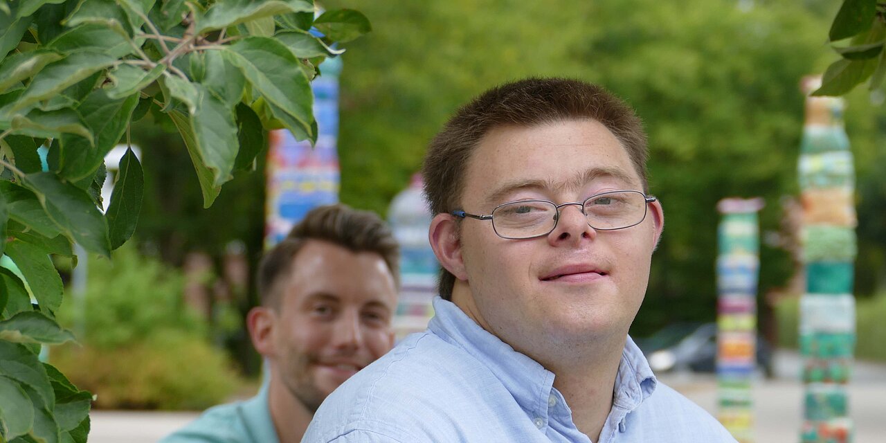 Ein junger Mann mit Downsyndrom lächelt in die Kamera, im Hintergrund ein Mitarbeiter