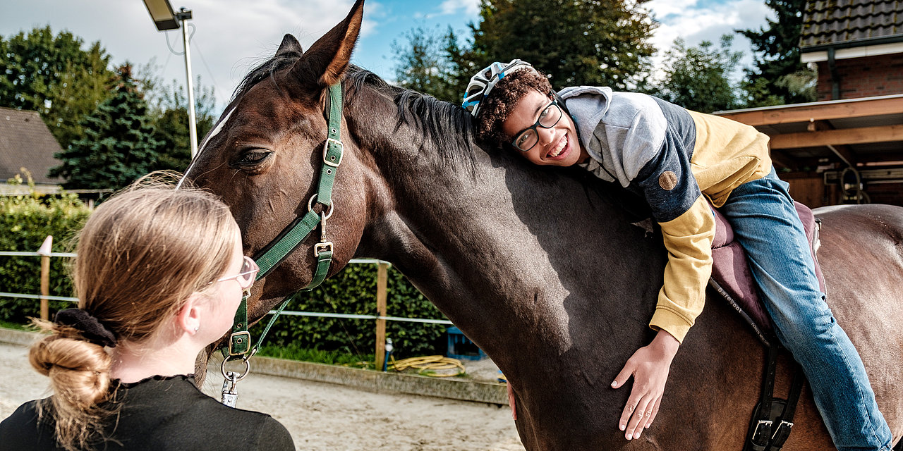 Ein Junge sitzt auf dem Rücken eines Pferdes und umarmt es strahlend, eine Frau hält das Pferd am Zaumzeug 