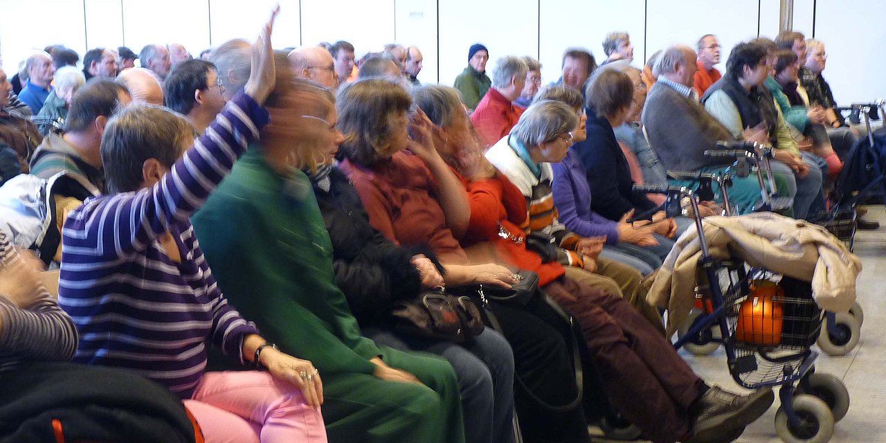 Viele Menschen sitzen in Stuhlreihen in einem Saal, einige von ihnen haben die Hand gehoben