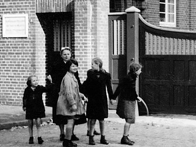 Ein historisches Schwarz-weiß-Foto zeigt eine Gruppe von Kindern vor dem geschlossenen Tür der Anstalten, Diakonissen begleiten die Kinder