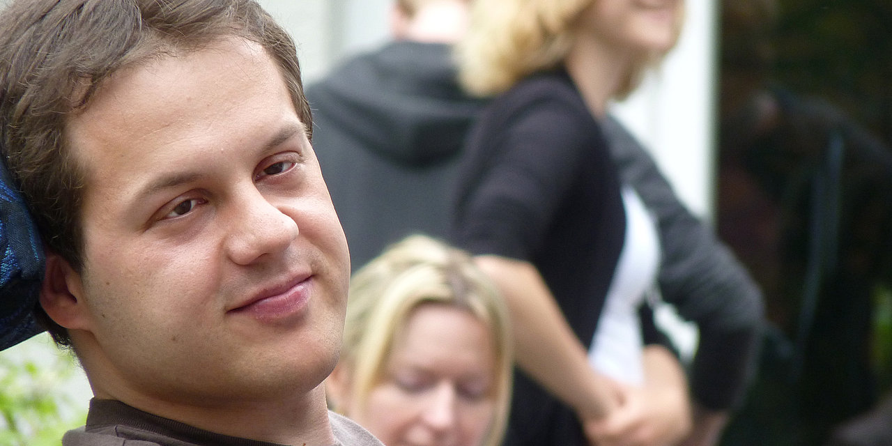 Ein Rollstuhlfahrer blickt verträumt in die Kamera, im Hintergrund sind junge Leute zu sehen