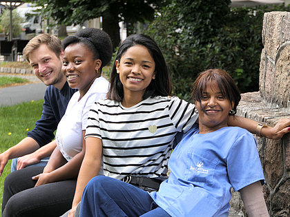 Vier junge Menschen unterschiedlicher Nation sitzen unter freiem Himmel beieinander und lächeln in die Kamera