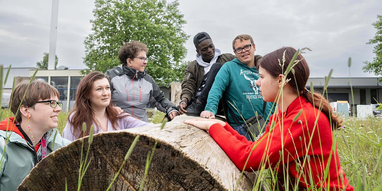 Sechs junge Menschen haben sich im Freien auf einer Wiese um einen liegenden Baumstamm versammelt