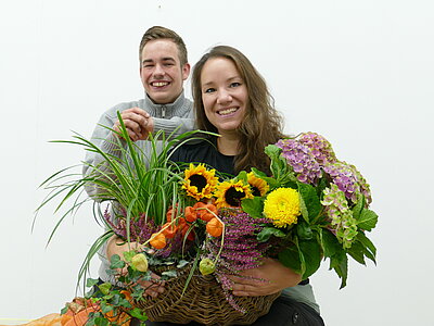 Eine junge Frau und ein junger Mann halten bunte Blumensträuße in der Hand und strahlen in die Kamera