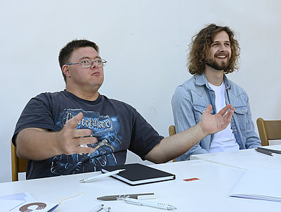 Zwei junge Männer sitzen am Tisch, schauen in die selbe Richtung und sind im Gespräche