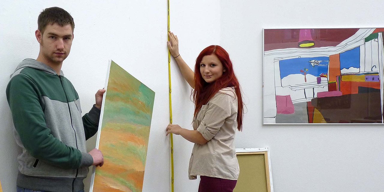 Zwei junge Menschen stehen im Atelier der Bildnerischen Werkstatt und halten Bilder in die Hand