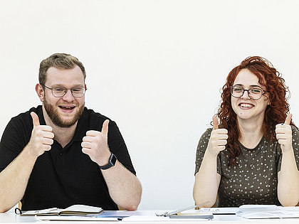 Drei junge Menschen sitzen an einem Tisch und blicken mit erhobenen Daumen in die Kamera