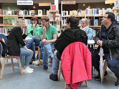 Mehrere junge Leute sitzen in einer Bücherei im Kreis und unterhalten sich