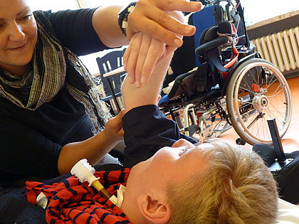 Eine Physiotherapeutin behandelt ein auf dem Rücken liegendes Kind, im Hintergrund steht ein Rollstuhl einer