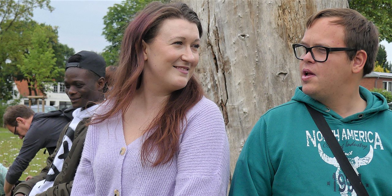 Eine junge Frau und ein junger Mann lehnen an einem Baumstamm und sind im Gespräch