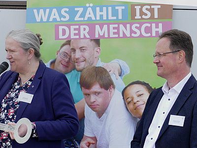 Sabine Ulrich und Thorsten Tillner von der Geschäftsführung blicken in einer Richtung, Frau Ulrich spricht in ein Mikrofon. Im Hintergrund ein Bild, dass eine Menschengruppe zeigt mit dem Slogan "Was zählt ist der Mensch"