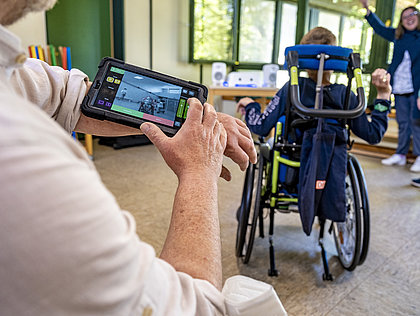Im Vordergrund hat ein Mitarbeiter ein Gerät mit Display um den Unterarm gebunden, im Hintergrund sitzt ein Mensch im Rollstuhl und wird von einer Frau animiert, die Arme zu heben.