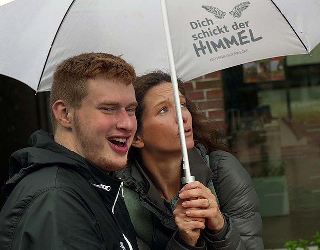 Ein junger Mann und eine junge Frau stehen unter einem Regenschirm mit der Aufschrift Dich schickt der Himmel