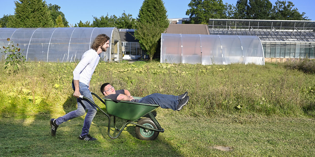 Ein junger Mann schiebt eine Schubkarre mit einem Bewohner darin über eine Wiese